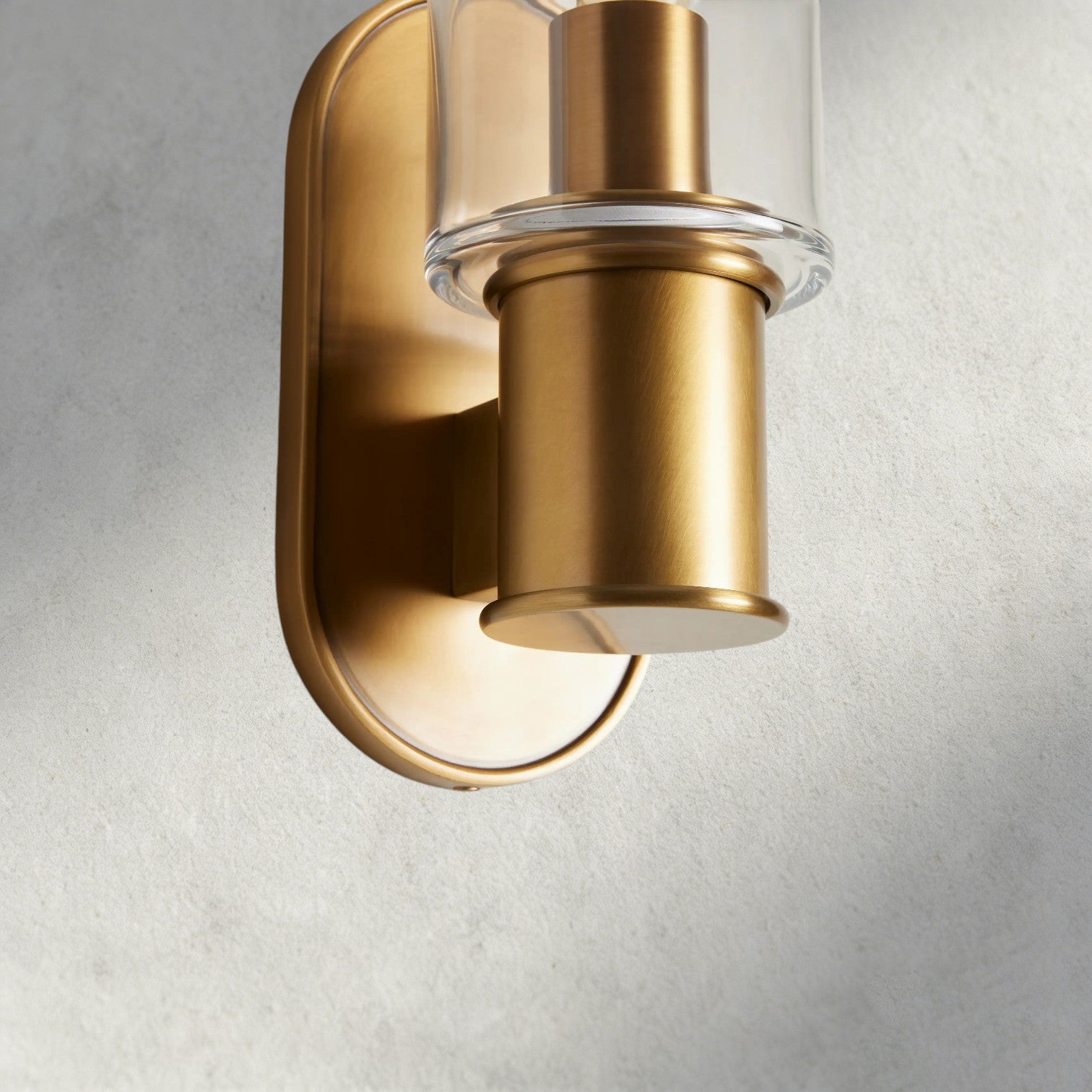 Lampthe Harlin Sconce in clear glass and brushed brass finish, featuring a modern oval backplate and sophisticated design.